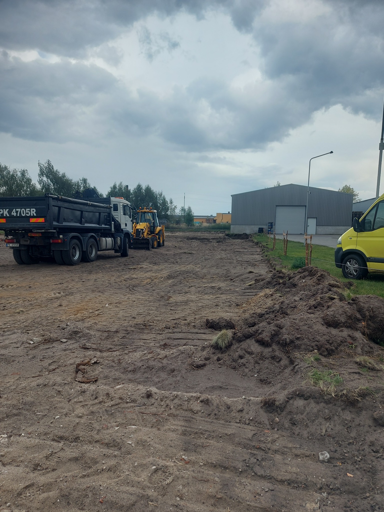 Przygotowanie terenu pod budowę: ciężarówka wyładowuje ziemię, a koparko-ładowarka wyrównuje teren, widoczne paliki wyznaczające granice.