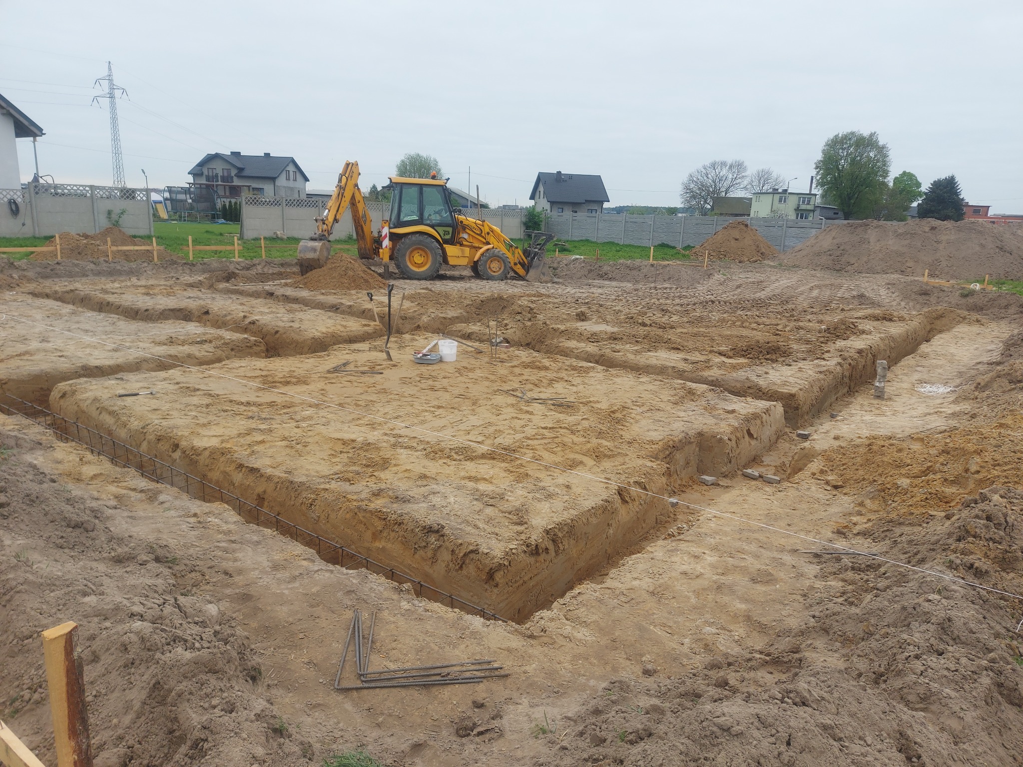Żółta koparko-ładowarka JCB przy wykopie pod fundamenty domu jednorodzinnego, widoczne zbrojenie w wykopie, teren przygotowany pod wylewkę.