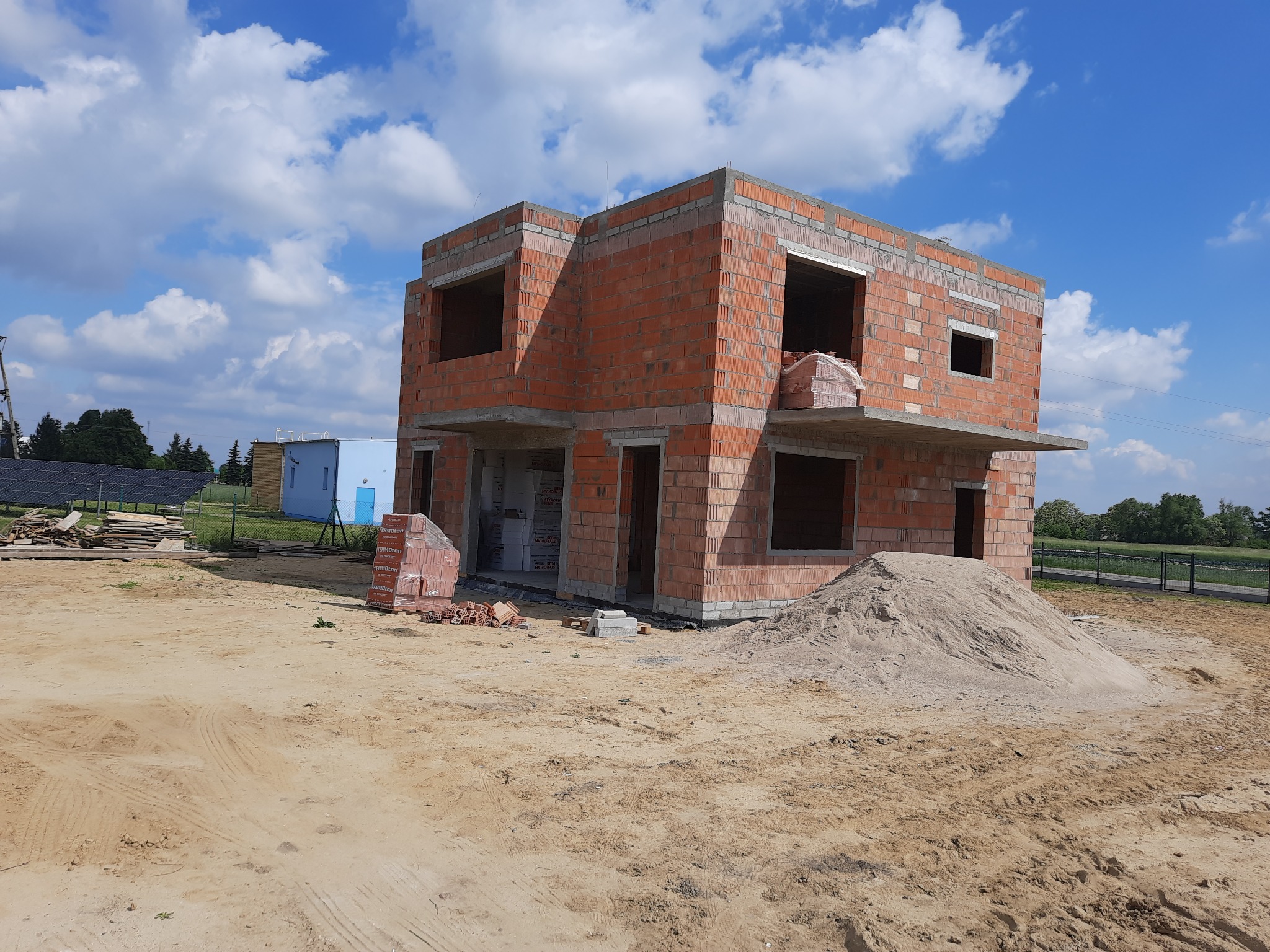 Dwukondygnacyjny budynek w stanie surowym otwartym, wzniesiony z czerwonej cegły kratówki, z widocznym stosem cegieł i hałdą piasku na placu budowy, przy słonecznej pogodzie.