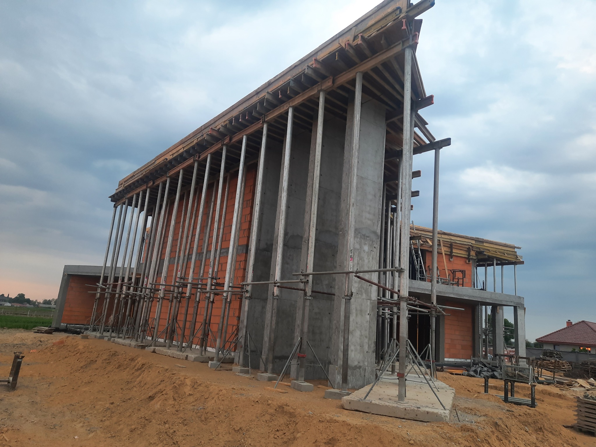 Budowa domu z czerwonej cegły w trakcie wznoszenia, widoczne betonowe filary i drewniane szalunki podtrzymujące konstrukcję, ziemny teren budowy.