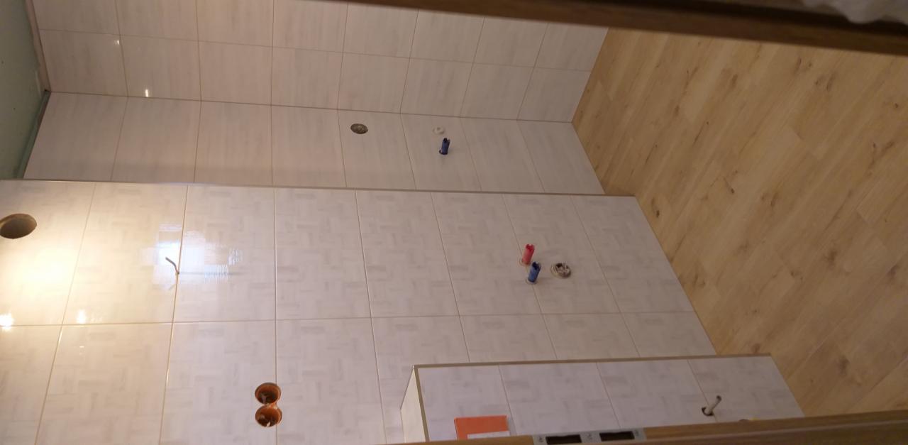 Ściana łazienki w trakcie kładzenia płytek, widoczne otwory na punkty elektryczne i rury, podłoga wyłożona panelami imitującymi drewno.