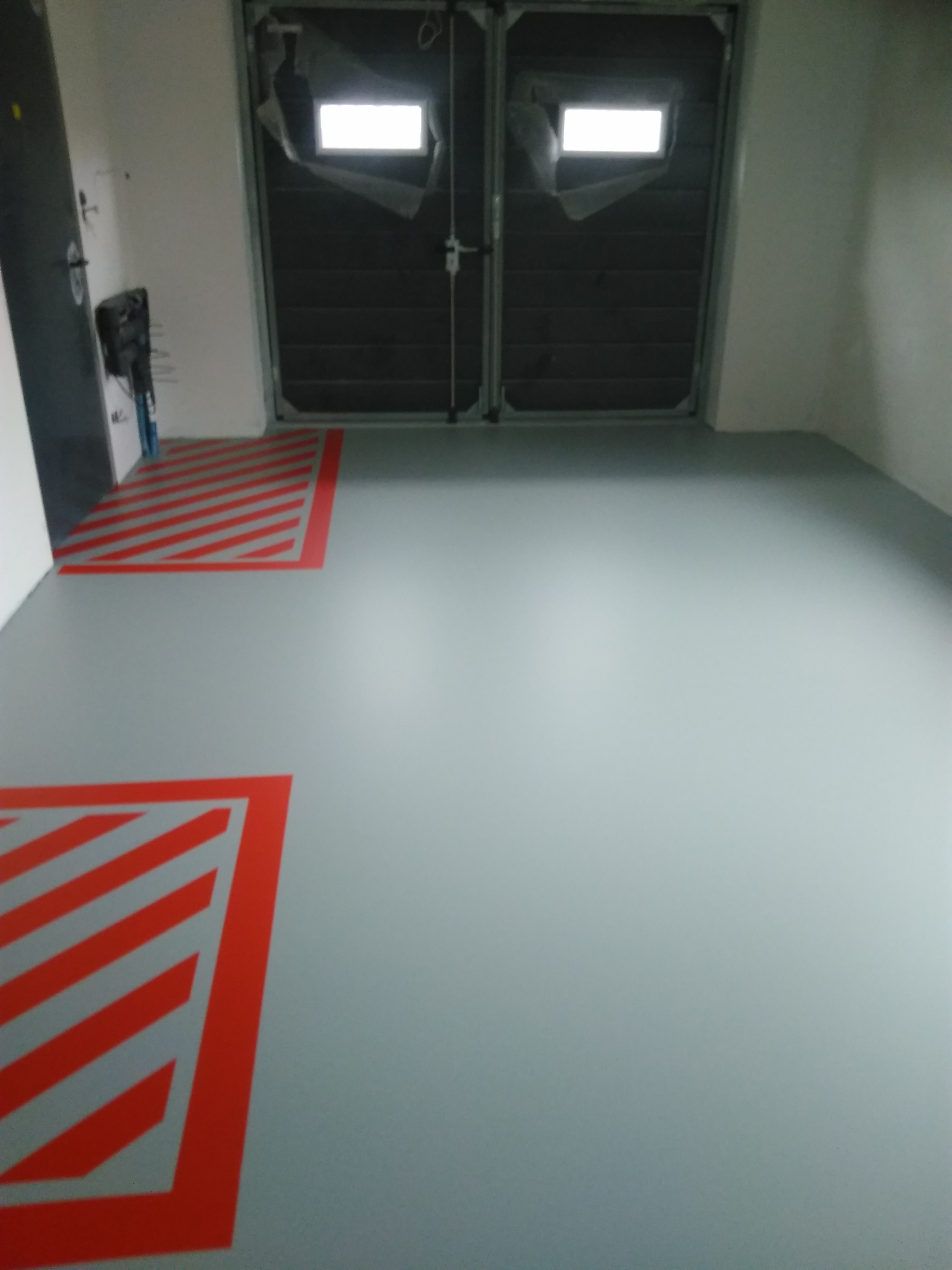 Szara, gładka podłoga w hali z czerwonymi pasami ostrzegawczymi przy ścianach, widoczne dwuskrzydłowe drzwi garażowe.