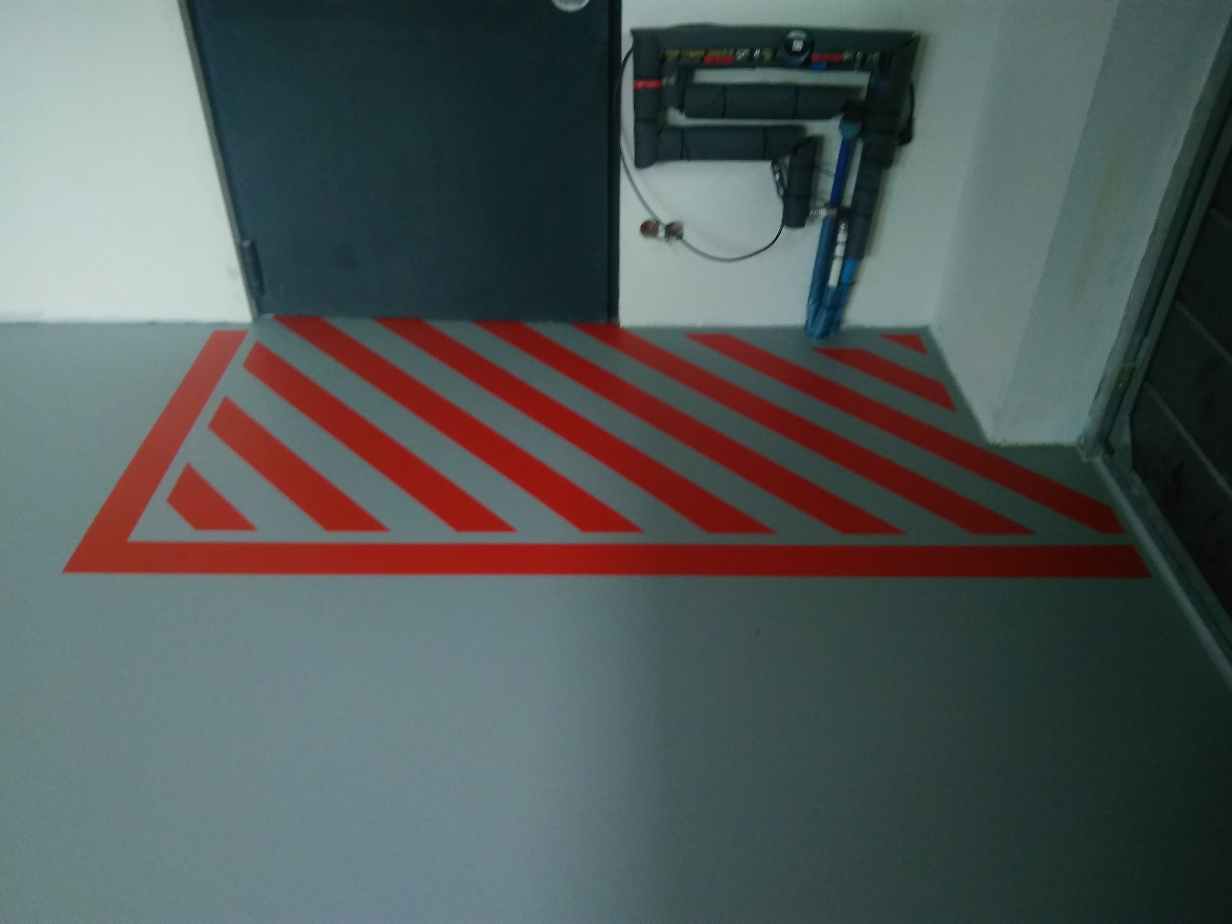 Szara posadzka z czerwonymi, ukośnymi pasami ostrzegawczymi przy ciemnych drzwiach i instalacji na ścianie.