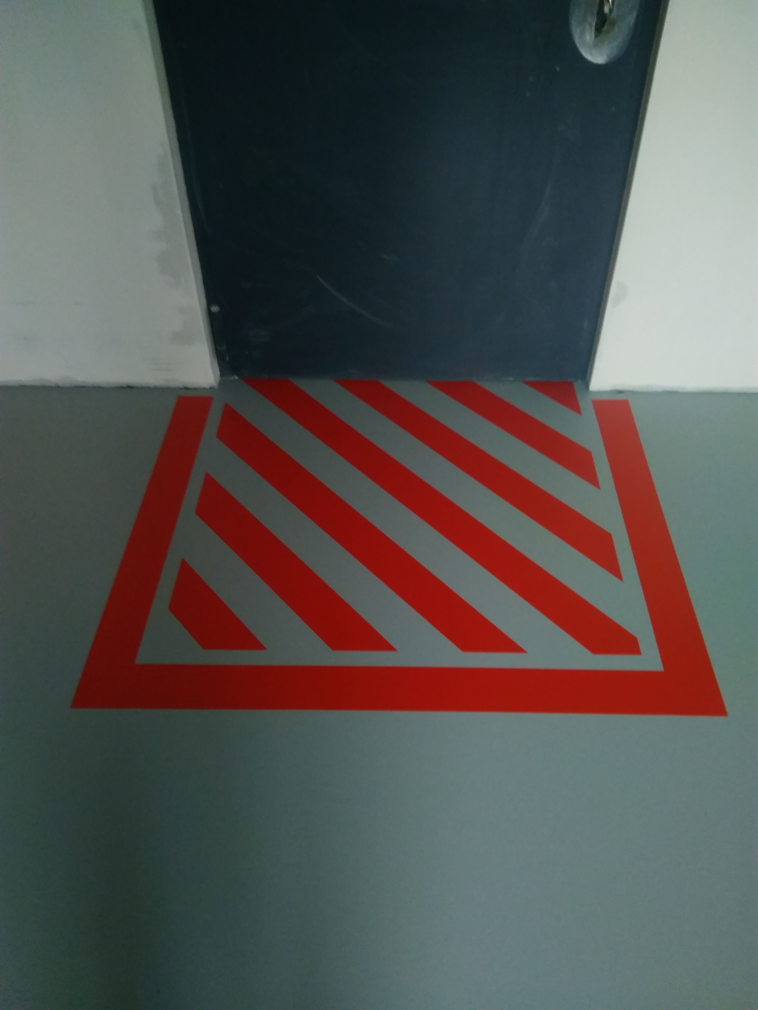 Szara posadzka żywiczna z czerwonymi, ukośnymi pasami ostrzegawczymi przy ciemnych drzwiach.
