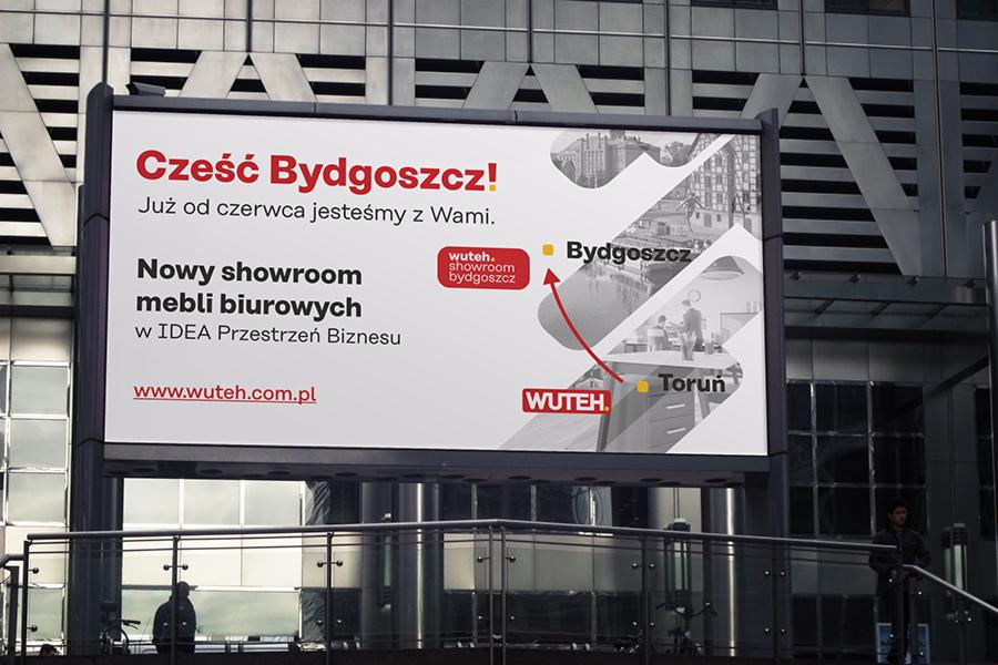 Billboard reklamujący nowy showroom mebli biurowych firmy WUTEH w Bydgoszczy, z mapą wskazującą lokalizacje w Bydgoszczy i Toruniu, umieszczony na tle nowoczesnego budynku.