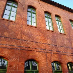 Czerwona ceglana ściana budynku z zielonymi, łukowatymi oknami, widoczna z dołu pod kątem, z cieniami drzew na fasadzie.