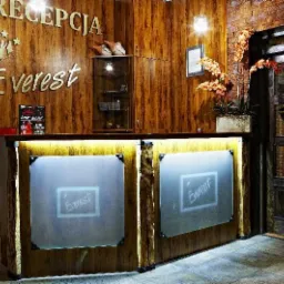 Recepcja hotelu Everest z drewnianą okładziną, podświetlanym blatem i logo na szklanych panelach, uzupełniona kwiatami i dekoracyjnymi elementami.