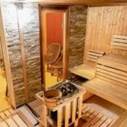 Wnętrze sauny z drewnianymi ławkami, piecem i kamienną ścianą. Przez otwarte drzwi widoczny wiklinowy fotel w przyległym pomieszczeniu.