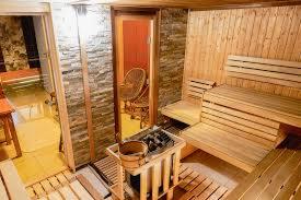 Wnętrze sauny z drewnianymi ławkami, piecem i kamienną ścianą. Przez otwarte drzwi widoczny wiklinowy fotel w przyległym pomieszczeniu.