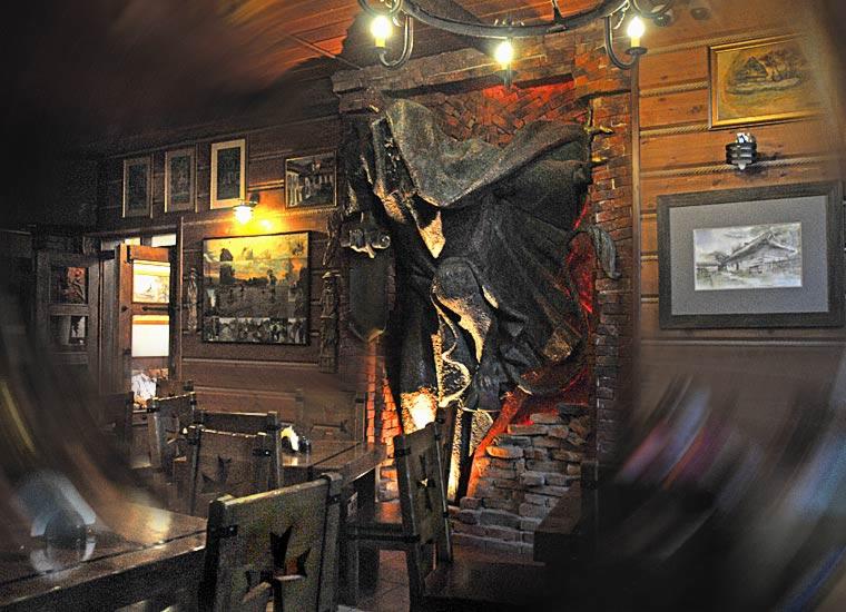 Rustykalne wnętrze restauracji z drewnianymi ścianami, stołami i krzesłami, oświetlone ciepłym światłem, z dekoracyjną rzeźbą na ceglanej ścianie i obrazami w ramach.