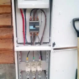 Otwarta skrzynka z bezpiecznikami i okablowaniem, widoczne trzy bezpieczniki topikowe, zamontowana przy drewnianej ścianie budynku.