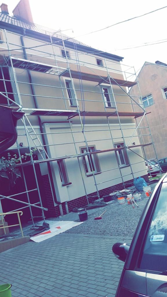 Dom w trakcie prac elewacyjnych, otoczony rusztowaniem, z widocznymi wiadrami z farbą i narzędziami malarskimi na ziemi.