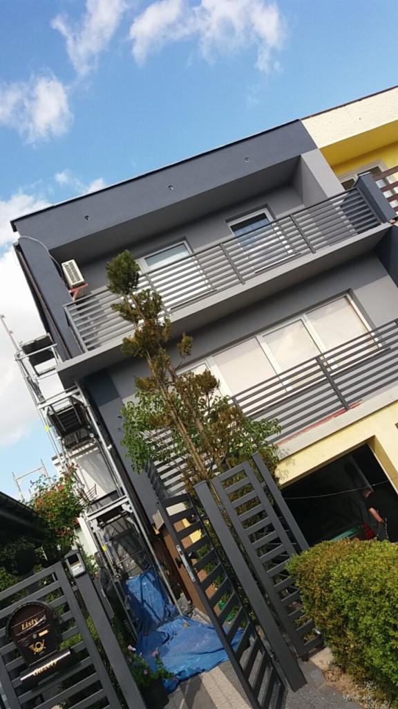 Nowoczesny dom z szarą elewacją i balkonami z metalowymi barierkami, widoczny z dołu, z rusztowaniem z boku i zielenią w ogrodzie.