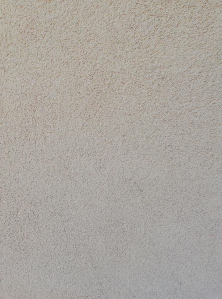 Jasnobeżowa elewacja o wyraźnej, ziarnistej fakturze, równomiernie oświetlona, z delikatnymi przebarwieniami w górnej części.