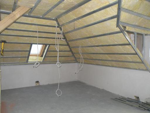 Poddasze w trakcie ocieplania: widoczna wełna mineralna między metalowymi profilami, okno dachowe, wystające kable elektryczne i żółty młotek na drewnianej belce.