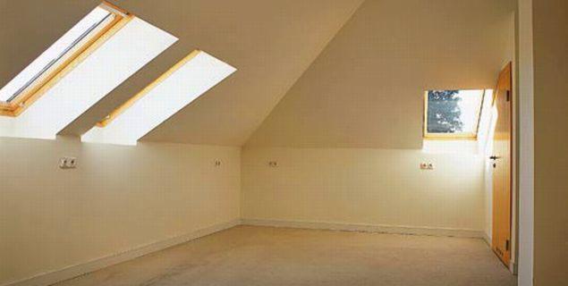 Poddasze z oknami dachowymi, ściany pomalowane na jasny kolor, widoczna instalacja elektryczna, drzwi po prawej stronie.