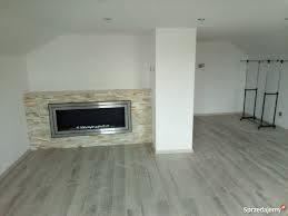 Poddasze po ociepleniu z wykończonym kominkiem elektrycznym w obudowie z jasnych płytek imitujących kamień, szara podłoga z paneli, białe ściany i kolumna, w tle widoczne składane wieszaki na ubrania.