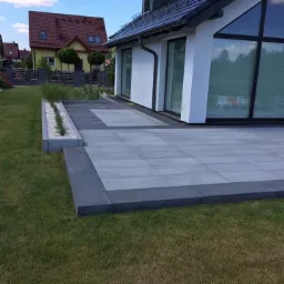Nowoczesny taras z szarej kostki brukowej ułożony przy białym domu z dużymi oknami, obrzeża z ciemniejszej kostki, widoczny trawnik i fragment ogrodu.