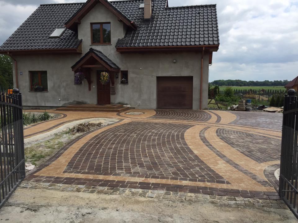 Ukończony podjazd z kostki brukowej w odcieniach brązu i beżu, ułożonej we wzór łuków prowadzących do domu z garażem, widok zza metalowej bramy.