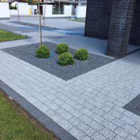 Nowoczesna nawierzchnia z szarej kostki brukowej ułożonej w regularny wzór, obrzeża z ciemniejszej kostki, centralnie kwadratowa rabata wypełniona szarym grysem i czterema kulistymi krzewami, w tle...
