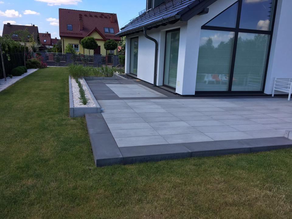Nowoczesny taras z szarej kostki brukowej ułożony przy białym domu z dużymi oknami, obrzeża z ciemniejszej kostki, widoczny trawnik i fragment ogrodu.
