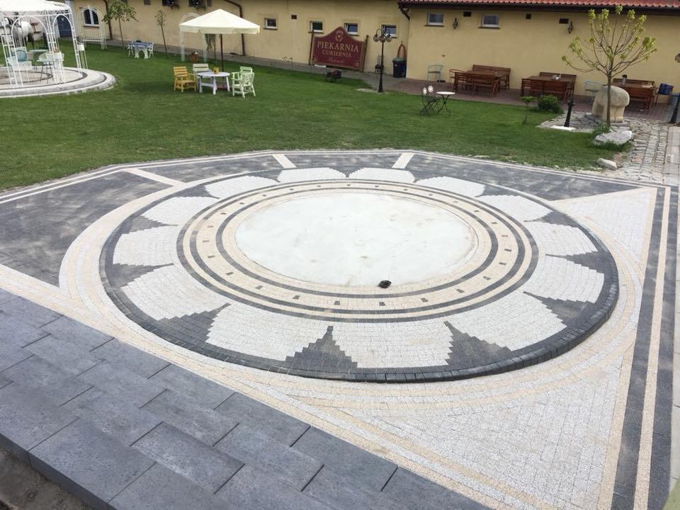 Okrągły plac z kostki granitowej ułożonej we wzór kwiatu, otoczony trawnikiem i budynkiem piekarni w tle.