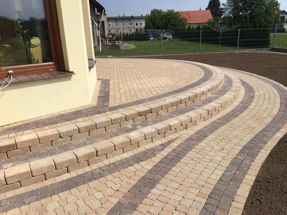 Schody i ścieżka z kostki brukowej ułożone w łukowaty wzór przy domu, z widocznym świeżo przygotowanym terenem pod trawnik w tle.