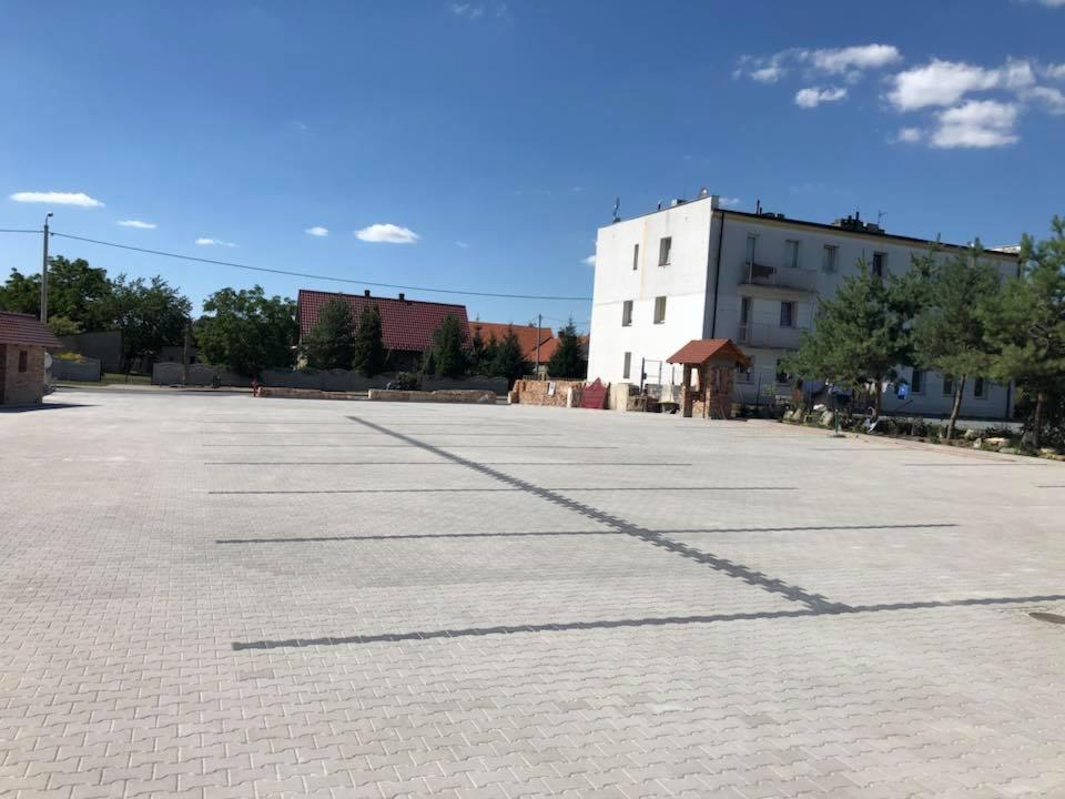 Rozległy parking wyłożony szarą kostką brukową w jodełkę, z widocznymi liniami parkingowymi w formie ciemniejszych pasów, budynek w tle i błękitne niebo.