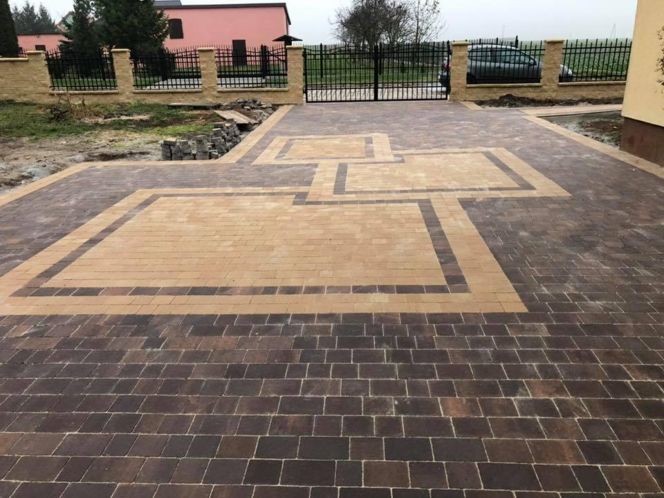 Ułożona nawierzchnia z kostki brukowej w dwóch odcieniach brązu i beżu, tworząca geometryczny wzór na podjeździe przed domem z czarną bramą w tle.