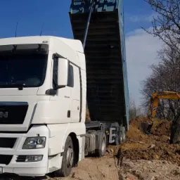 Biały samochód ciężarowy MAN z podniesioną naczepą wysypuje ziemię, w tle żółta koparka Liebherr i drzewa bez liści.