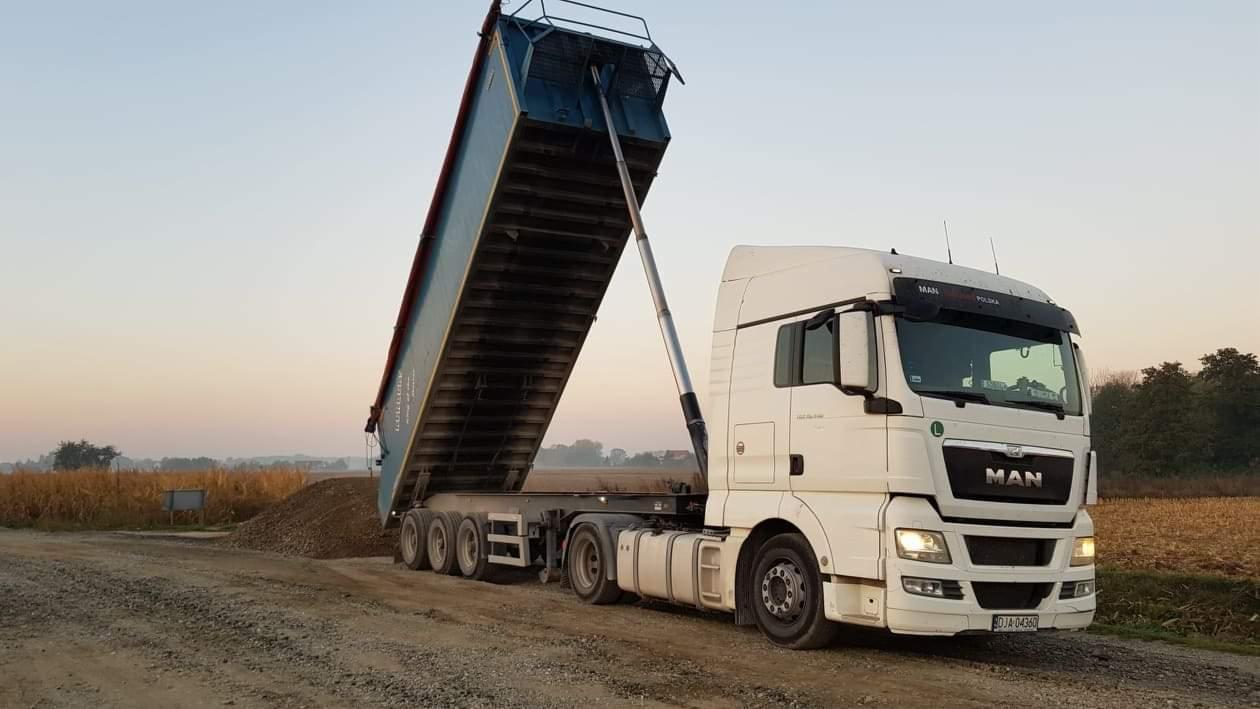 Biała ciężarówka MAN z podniesioną naczepą wysypuje żwir na polną drogę przy zachodzącym słońcu.