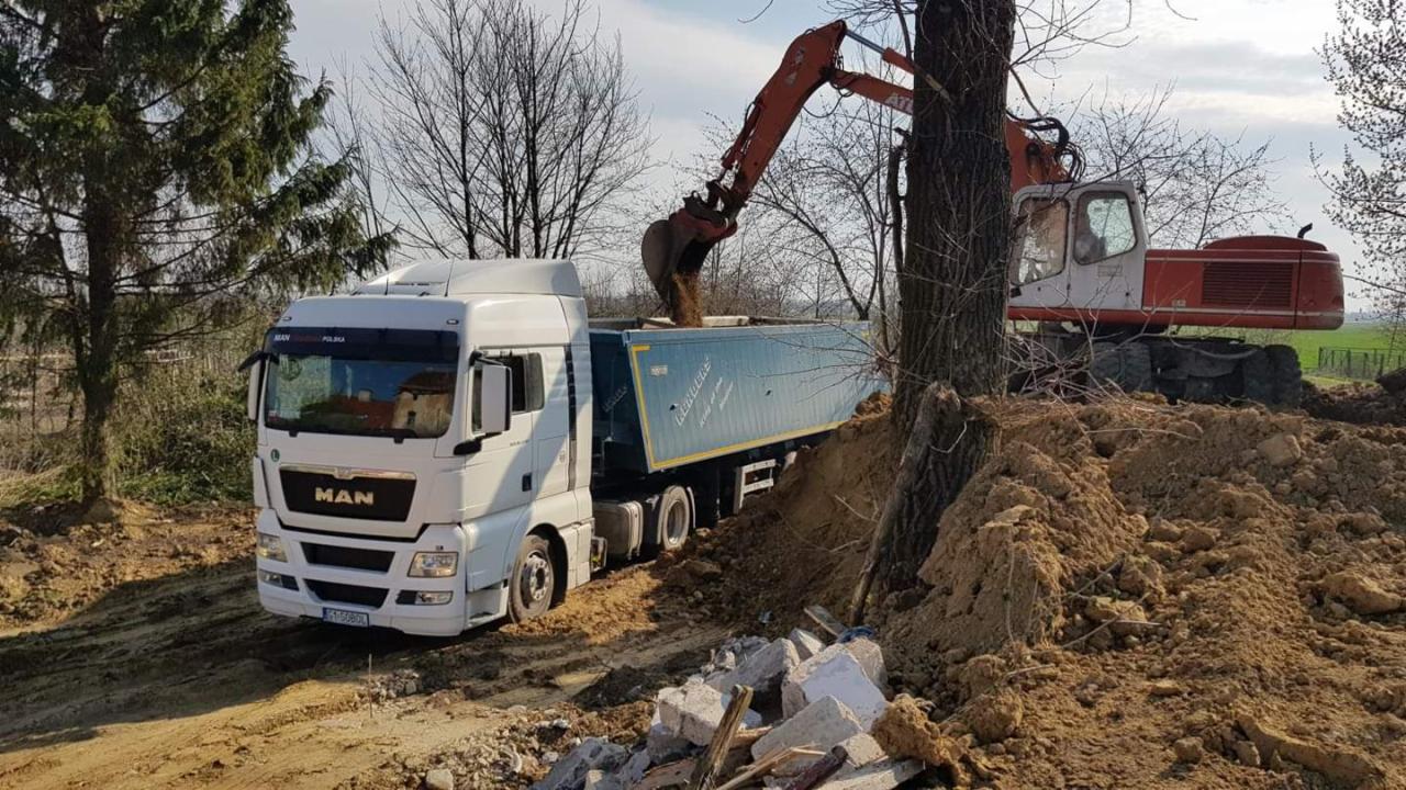 Ciężarówka MAN z naczepą, na którą koparka nabiera ziemię, w tle drzewa i pole.