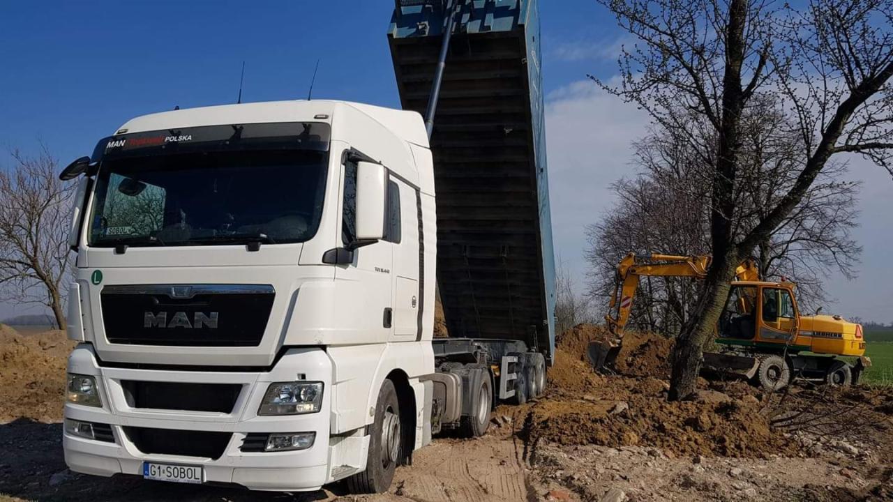 Biały samochód ciężarowy MAN z podniesioną naczepą wysypuje ziemię, w tle żółta koparka Liebherr i drzewa bez liści.