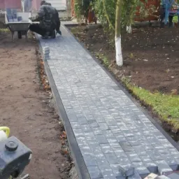 Układanie szarej kostki brukowej na ścieżce w ogrodzie, widoczna ubijarka wibrująca i pracownik z taczką kostki w tle.