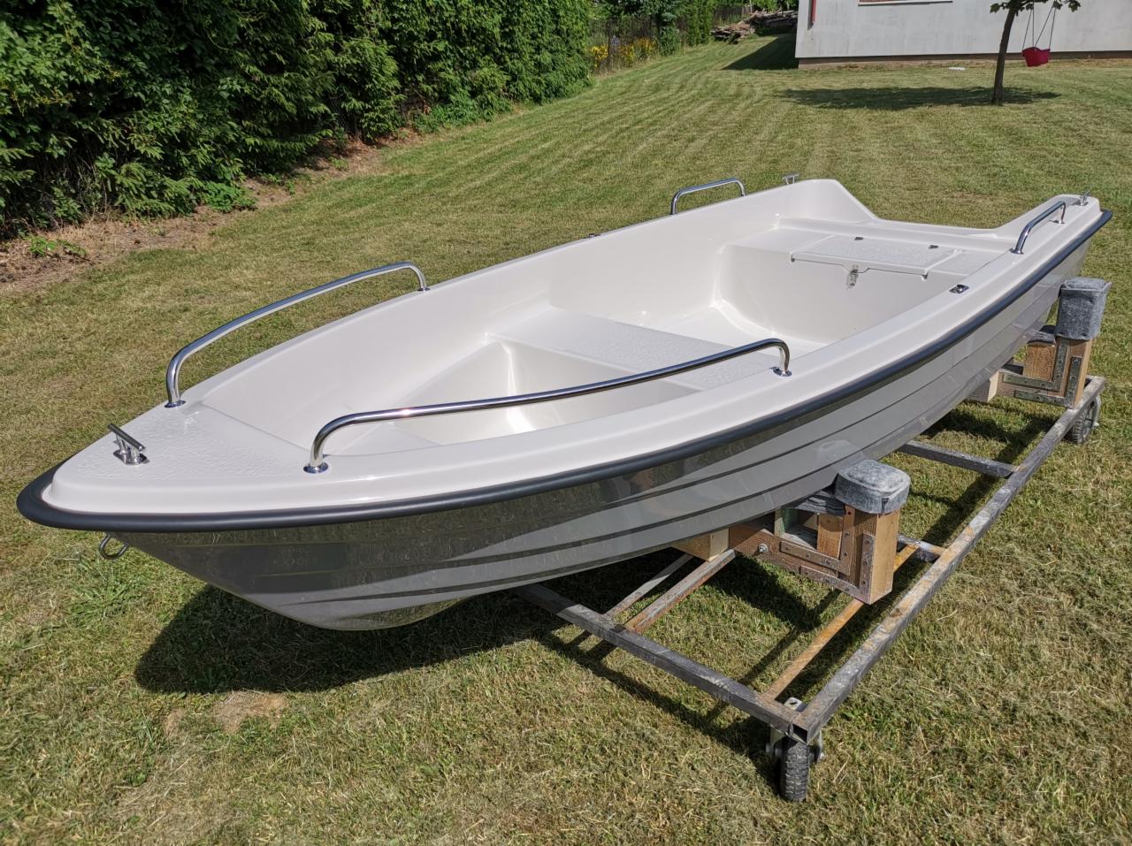 Biała łódka z relingami na metalowym wózku na trawniku przed domem.