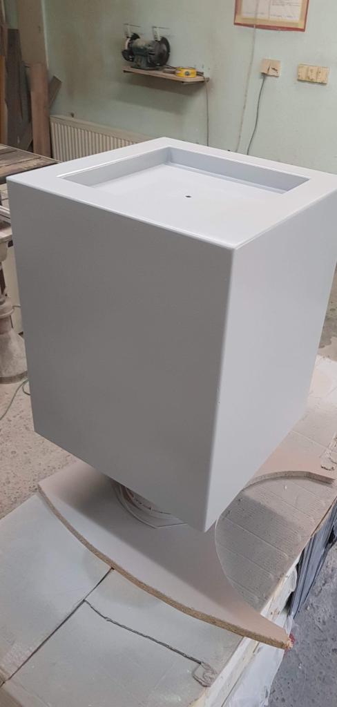 Szary, kwadratowy zbiornik z otworem w górnej części, stojący na obrotowej podstawie w warsztacie, prawdopodobnie w trakcie malowania lub wykańczania.