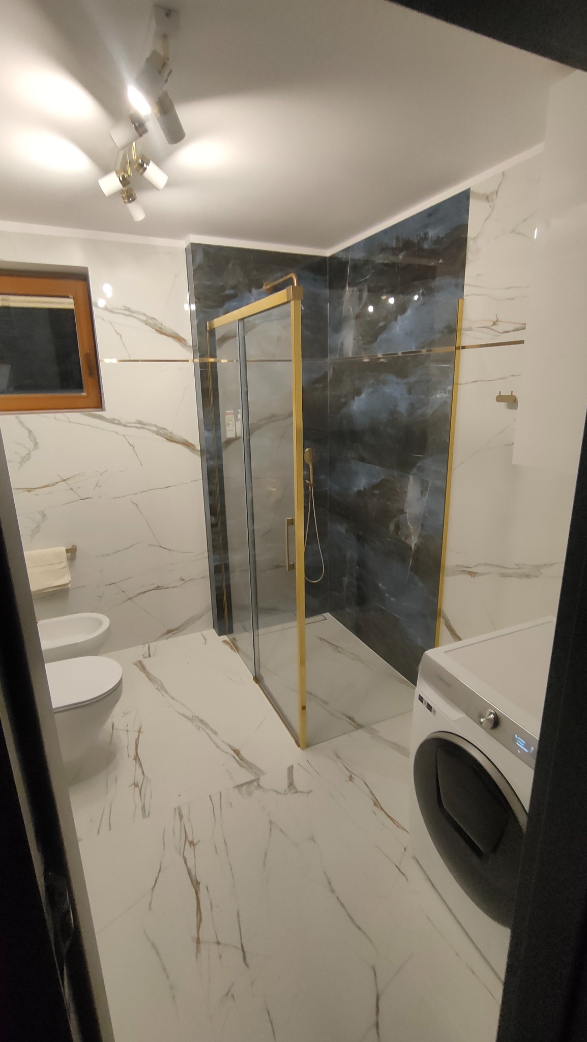 Nowoczesna łazienka z kabiną prysznicową w złotej ramie, marmurowymi płytkami na ścianach i podłodze, wiszącą toaletą i pralką.