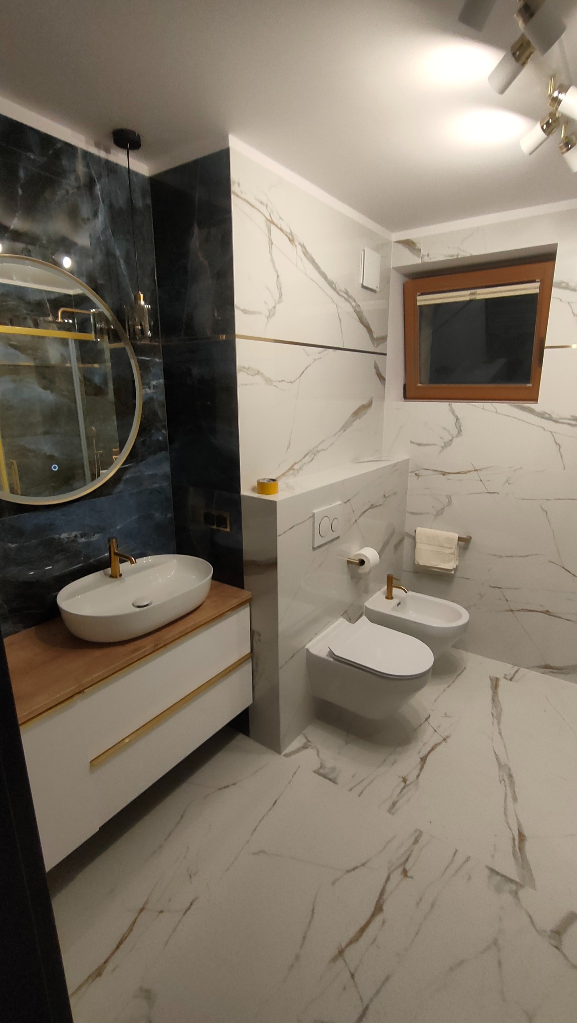 Elegancka łazienka z marmurowymi płytkami, okrągłym lustrem z podświetleniem LED, białą umywalką na drewnianym blacie z złotymi detalami, wiszącą toaletą i bidetem.