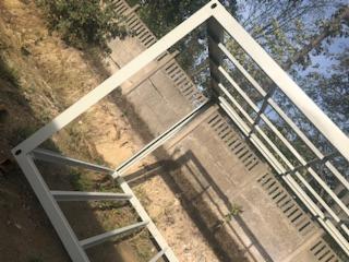 Metalowa konstrukcja szkieletowa, prawdopodobnie spawana, pomalowana na biało, leżąca na betonowej powierzchni z widocznymi elementami roślinności i ogrodzenia w tle.