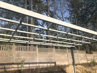 Konstrukcja metalowa z profili zamkniętych, pomalowana na biało, tworząca zadaszenie, widoczna na tle betonowego ogrodzenia i drzew.