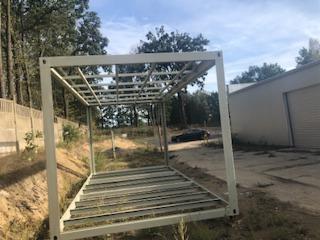 Metalowa konstrukcja szkieletowa, prawdopodobnie pod budynek lub pawilon, na zewnątrz, w tle widoczne drzewa i budynek magazynowy.