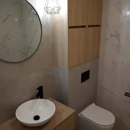 Nowoczesna łazienka z okrągłym lustrem, wiszącą szafką z drewnianym fornirem, białą umywalką nablatową z czarną armaturą i podwieszanym WC, oświetlona lampą wiszącą z geometrycznym kloszem.