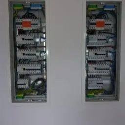 Dwa panele elektryczne z widocznymi zabezpieczeniami i okablowaniem, zamontowane w białej ścianie, z lekkim cieniem na lewym panelu.