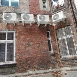 Cztery klimatyzatory zamontowane na zewnątrz budynku z czerwonej cegły, widoczne okna z kratami i rynna spustowa.