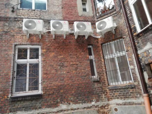 Cztery klimatyzatory zamontowane na zewnątrz budynku z czerwonej cegły, widoczne okna z kratami i rynna spustowa.