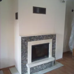 Obudowa kominka z kamienia łupanego w odcieniach szarości i beżu, z jasnym, kremowym obramowaniem, na tle jasnych ścian i drewnianej podłogi.