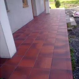 Wyłożony czerwoną terakotą taras przy domu z widocznymi schodami wejściowymi i fragmentem ogrodu.