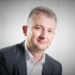 Portret biznesowy mężczyzny w szarej marynarce i białej koszuli, uśmiechającego się do kamery na jasnym tle. Widoczny fragment ramienia i tors.