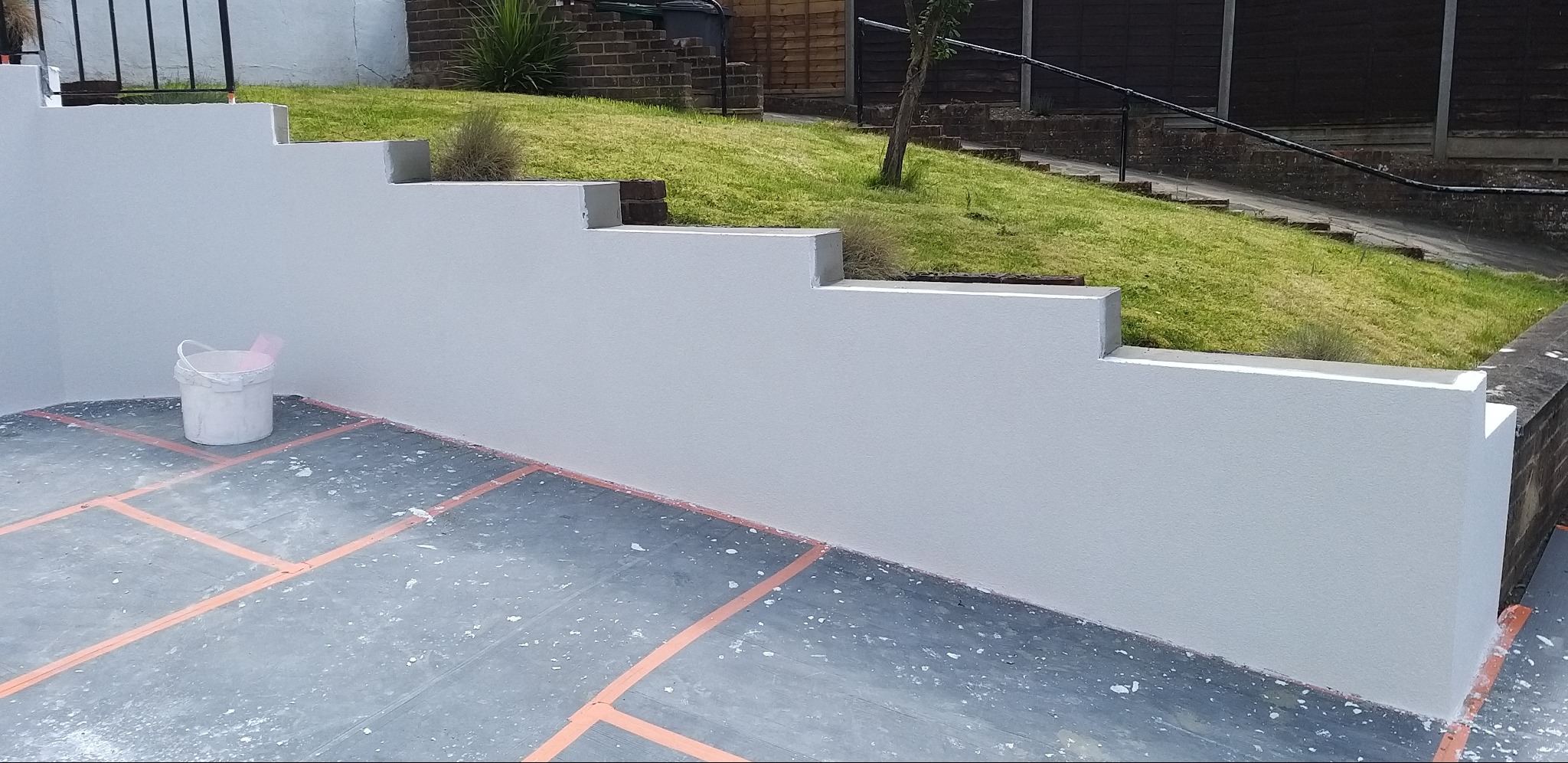 Świeżo pomalowany, schodkowy mur oporowy w ogrodzie, z widocznym wiadrem z farbą i zabezpieczonym podłożem.