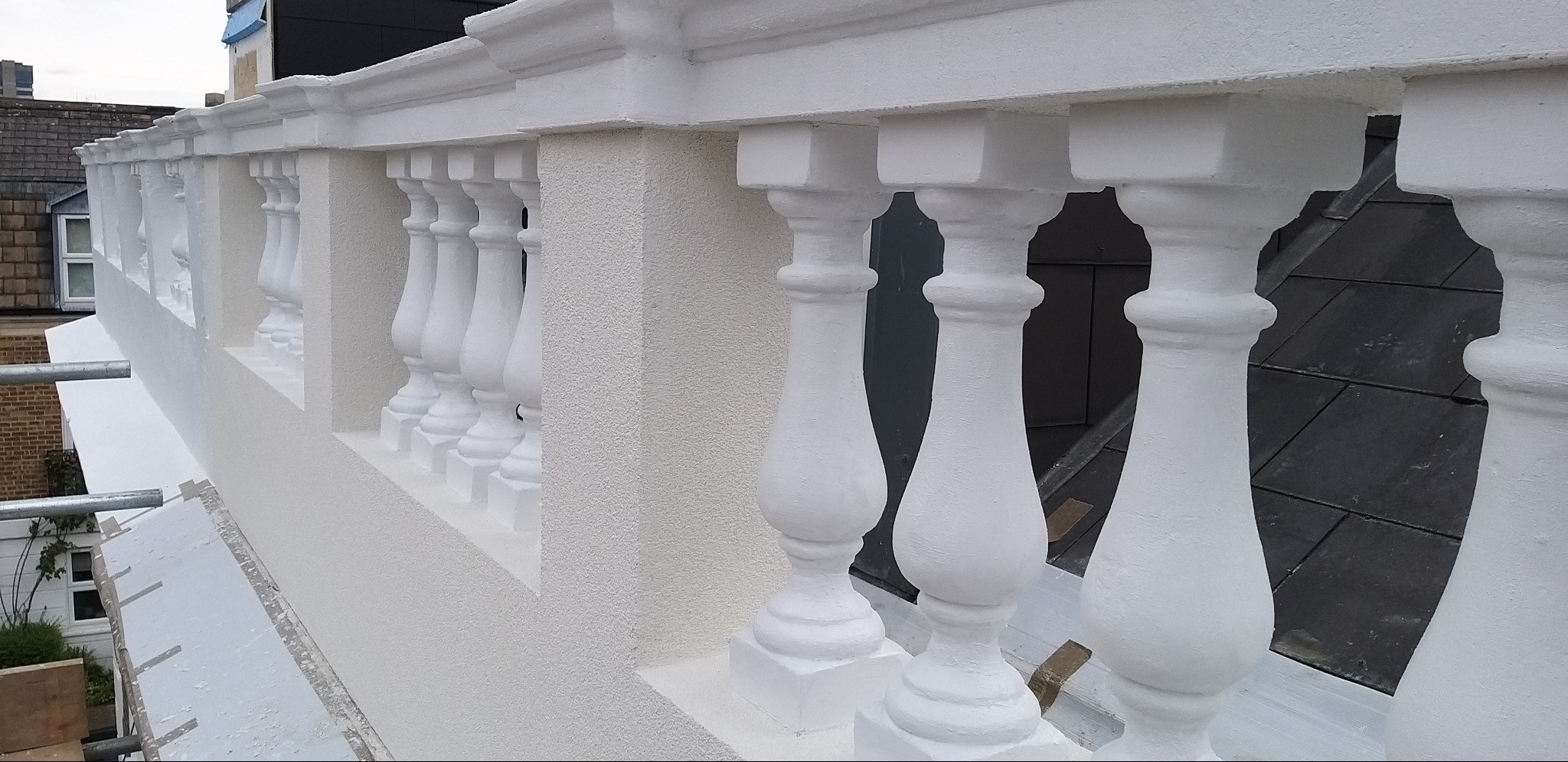 Biała balustrada balkonowa z dekoracyjnymi tralkami i teksturowaną ścianą, widok z góry, fragment rusztowania w lewym dolnym rogu.